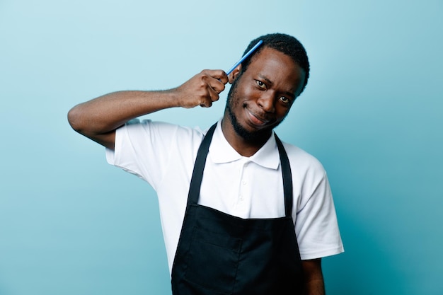 Smiling combing hair with comb young african american barber in uniform isolated on blue background