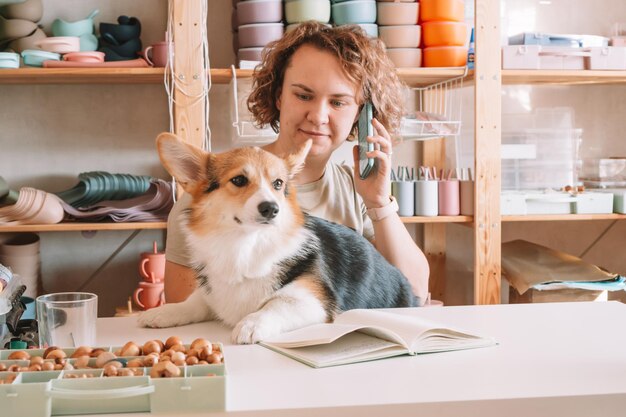 Smiling concentrated businesswoman talking smartphone holding cute dog corgi in workshop Silicone baby dishes shop