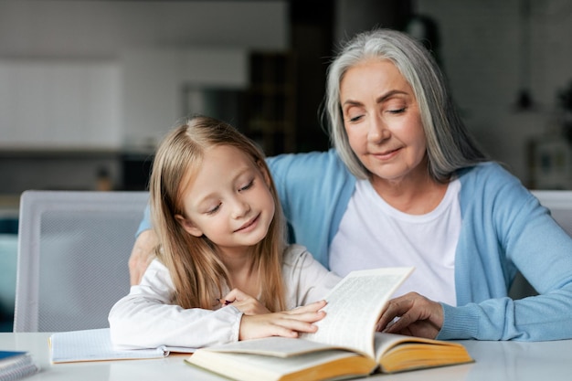 은퇴한 할머니와 함께 웃고 있는 집중된 똑똑한 유럽 어린 소녀는 책을 읽고 공부를 즐긴다