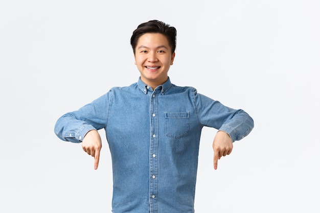 Smiling confident asian man satisfied with his stomatology clinic, recommend doctor, showing braces proud, pointing fingers down satisfied, standing white background