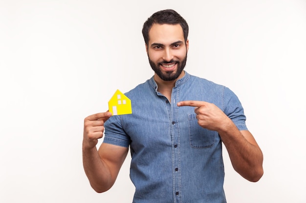 Smiling confident bearded man in blue shirt pointing finger at little paper house he holding in hand, real estate agency, accommodation. Indoor studio shot isolated on white background