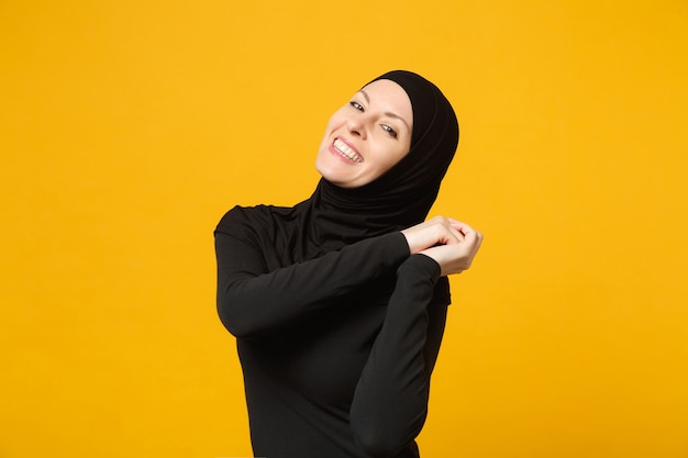 Smiling confident beautiful young arabian muslim woman in hijab black clothes posing isolated on yellow wall, portrait. People religious Islam lifestyle concept. 