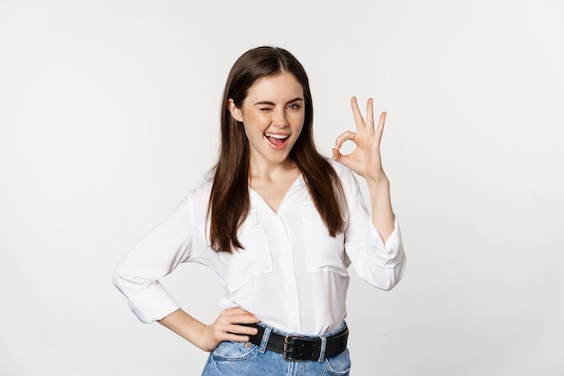 Smiling confident business woman winking showing okay ok sign excellent no problem gesture standing ...