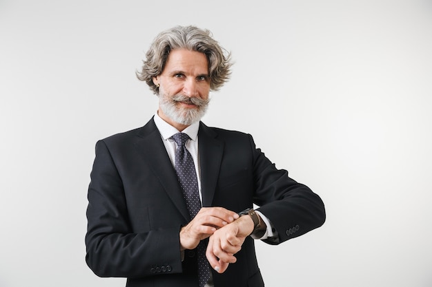 Smiling confident mature businessman wearing suit standing isolated over white wall, checking time