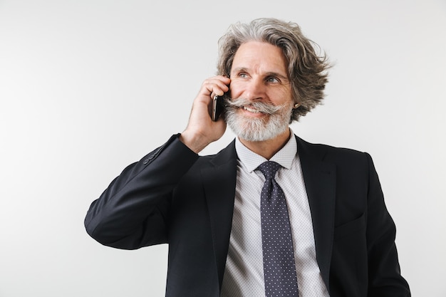 Smiling confident mature businessman wearing suit standing isolated over white wall, talking on mobile phone