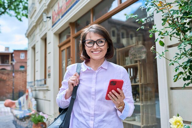 Smiling confident mature female looking at camera outdoor Positive beautiful middleaged female with smartphone on street of sunny summer city Age beauty vacation walk lifestyle concept