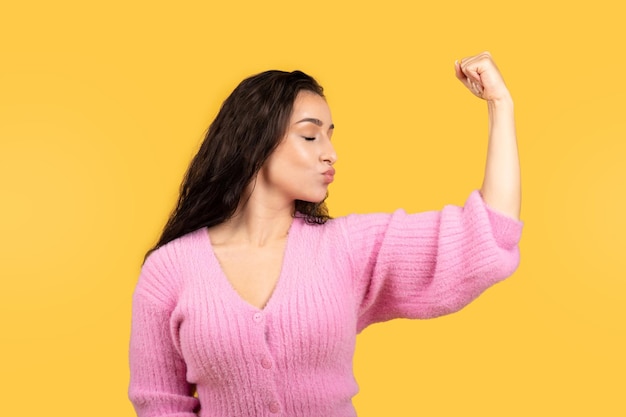 Smiling confident millennial middle eastern lady kissing biceps on hand isolated on yellow studio