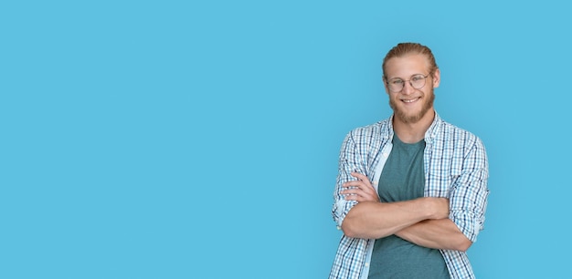 Smiling confident young bearded handsome man hipster wear shirt glasses looking at camera millennial handsome casual guy student arms crossed isolated on blue studio background portrait copy space