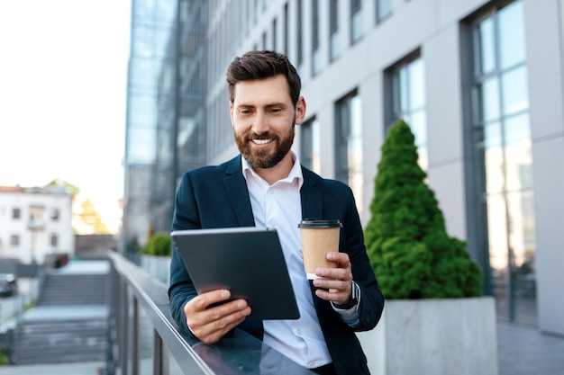 スーツを着たひげを生やした笑顔の自信に満ちた若い白人男性は、タブレットでビデオ通話を行い、持ち帰り用のコーヒーを飲みます