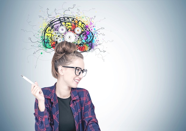 smiling confident young hipster woman wearing a checkered shirt, a black t shirt and glasses holding a pen and looking sideways. a gray wall background with a cog brain sketch on it. mock up