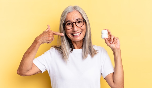 Smiling confidently pointing to own broad smile, positive, relaxed, satisfied attitude and holding a pills bottle