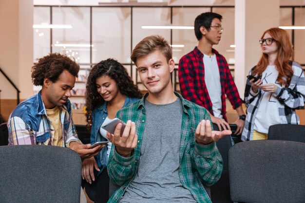 Smiling confused young man sitting and using mobile phone whie his friends talking