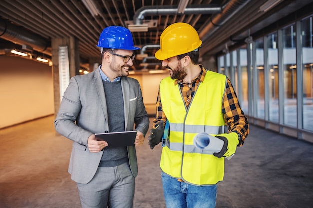 Smiling construction worker in working wear holding blueprints and talking with main architect about realization of new project.