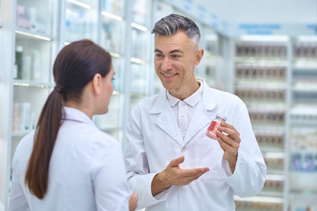 Sorridente e contento farmacista maschio dai capelli grigi che mostra una bottiglia di medicinale alla sua collega dai capelli scuri