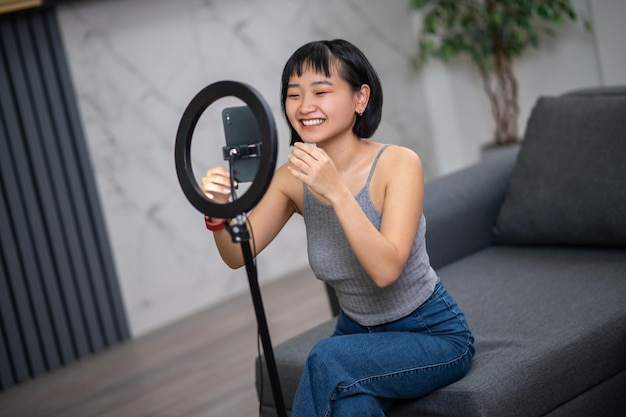 Smiling contented vlogger seated on the sofa before the smartphone mounted on the tripod stand