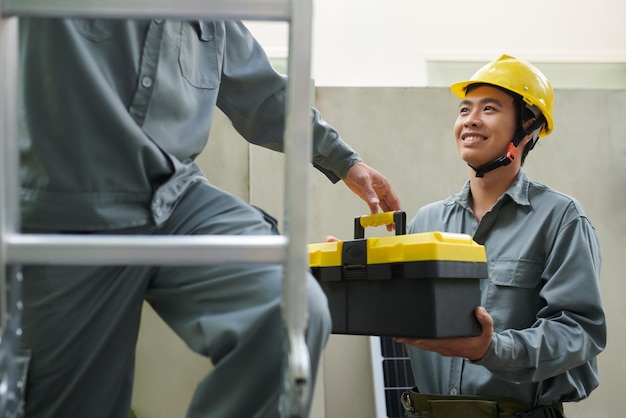 Smiling contractor giving heavy toolbox to coworker