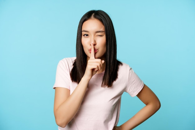 Sorridente ragazza asiatica civettuola che zittisce facendo silenzio tabù gesto strizzando l'occhio e facendo segno di silenzio pre...