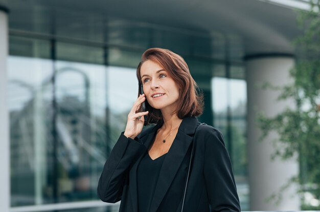 Photo smiling corporate person with ginger hair in black formal clothes talking on a mobile phone outdoors