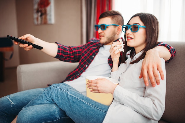 Smiling couple in 3D glasses sitting on couch and watch tv with popcorn at home, man with remote control in hand