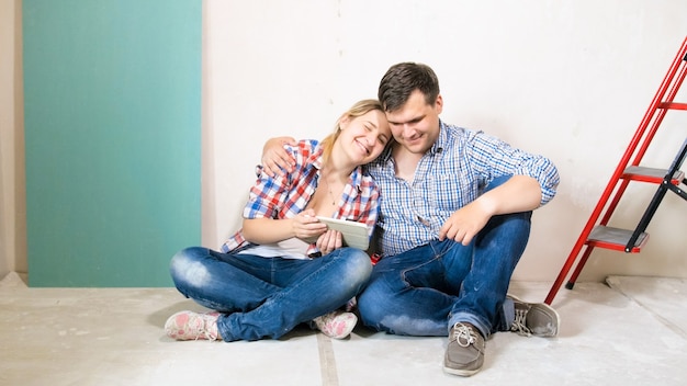 Smiling couple in home under renovation sitting on floor and holding digital tablet computer.