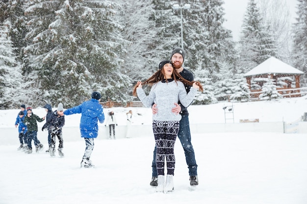 Улыбающаяся пара в коньках, обнимая на открытом воздухе со снегом на фоне