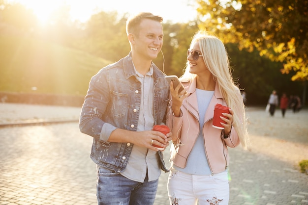 Smiling couple in love walking in autumn park, holding hands. Listen to music on headphones and drink coffee to go.