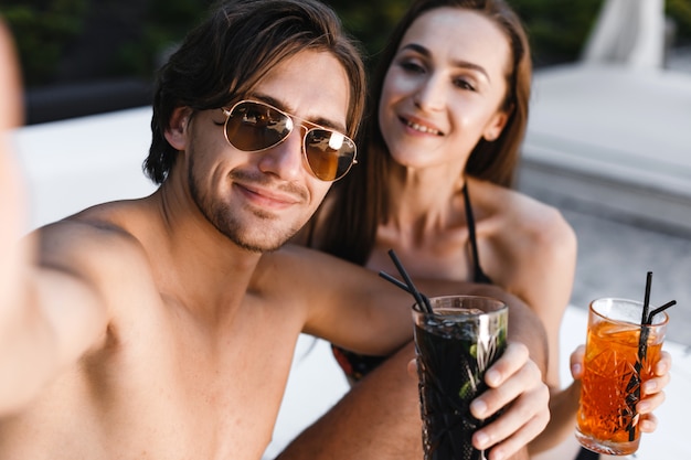 Smiling couple making selfie and holding cocktails at the beach