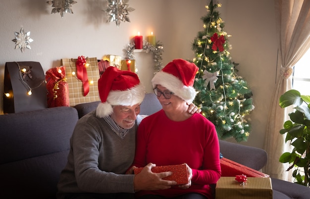 A smiling couple of man and woman in Santa's hats opening a Christmas present and are surprised by the content. Christmas tree and gifts for the family around them