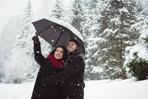 Coppia sorridente sotto l'ombrello che punta a vista nella foresta durante la nevicata