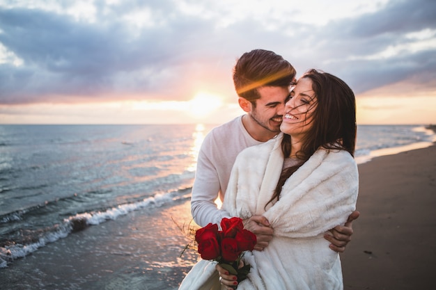 Smiling couple walking on the beach with a bouquet of roses at sunset