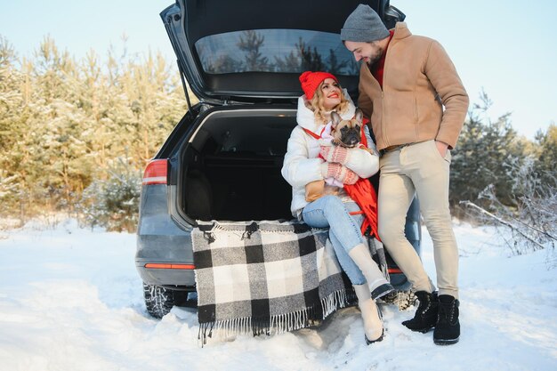 Smiling couple with dog sitting in open SUV car trunk in snowy forest Enjoying each other in active winter holidays