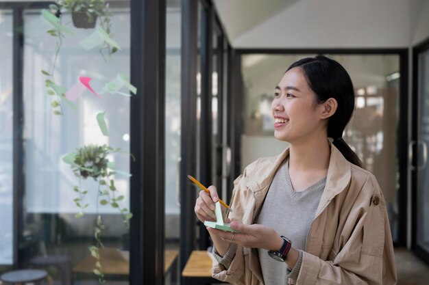 Smiling creative woman reading sticky notes on glass wall in modern workplace