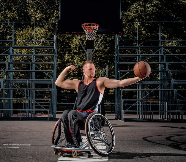 Smiling cripple basketball player in wheelchair holds a ball on open gaming ground.