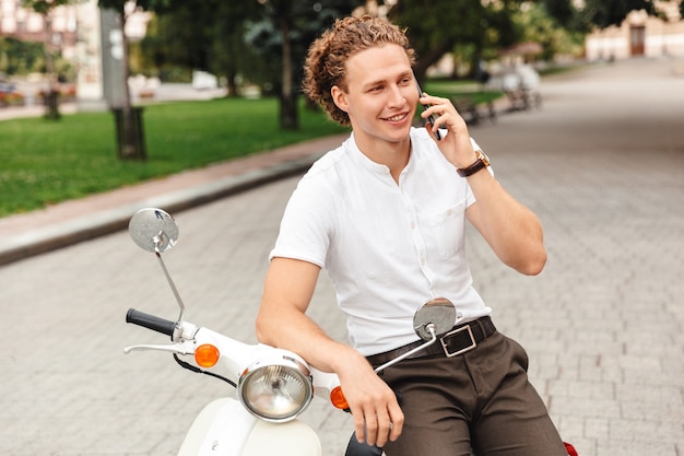 Smiling curly business man talking by smartphone and looking away while sitting on scooter outdoors