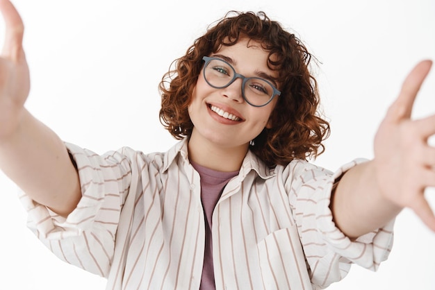 Smiling curly girl in glasses holding camera with stretch out hands video calling with tablet or phone reaching for hug standing against white background