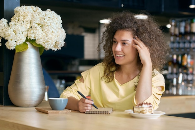 Foto ragazza riccia sorridente che si siede sul tavolo di legno che si tocca il viso che beve caffè