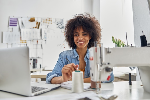 현대적인 노트북을 들고 웃고 있는 곱슬머리의 아프리카계 미국인 여성이 직장에 앉아 있습니다. 스튜디오의 재봉틀에서 새 옷 모델 작업을 하는 여성 재단사
