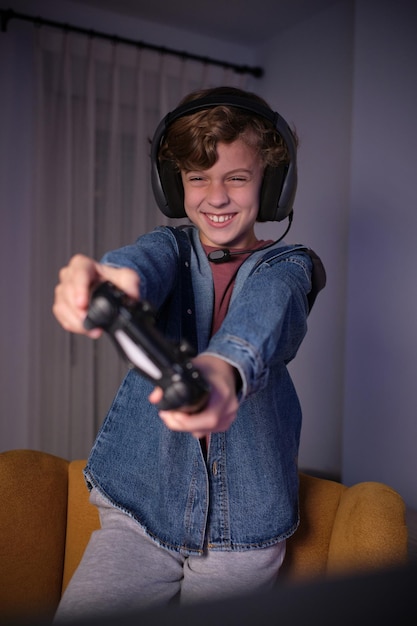 smiling curly haired preteen kid in headphones and casual clothes standing on armchair while playing videogame with joystick