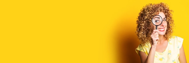 Photo smiling curly young woman looking through a magnifying glass on a yellow background. banner.