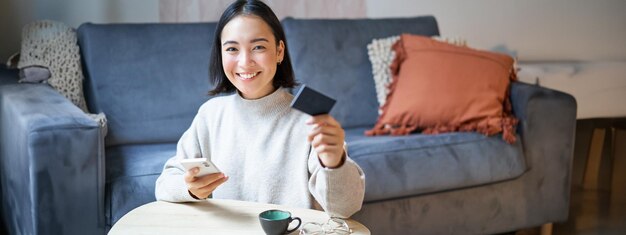 クレジット カードとスマートフォンを使用して、携帯電話を保持してオンラインで請求書を支払う笑顔のかわいいアジアの女性