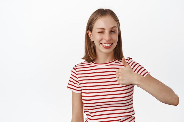 Smiling cute blond woman encourage you, showing thumb up and winking happy, approve, praise good choice, compliment you, white wall