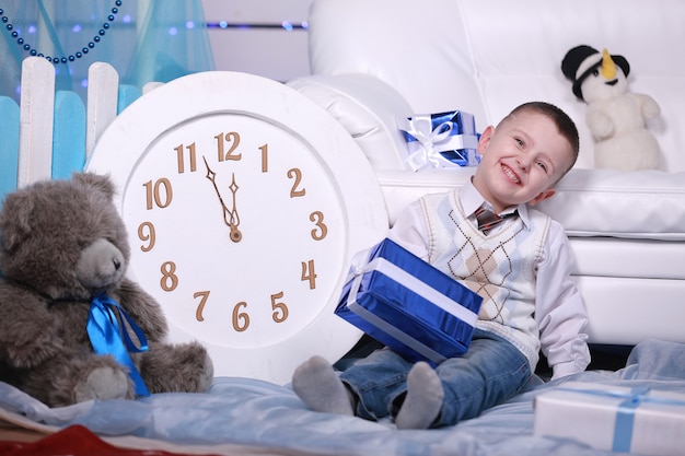 Foto ragazzo sveglio sorridente che tiene il suo presente durante il periodo natalizio. grande orologio bianco e orsacchiotto sul muro