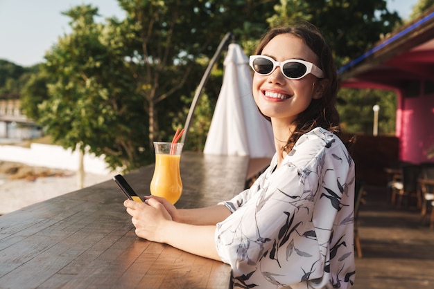 Smiling cute cheerful young beautiful woman at the beach drinking juice cocktail in cafe wearing sunglasses using mobile phone.