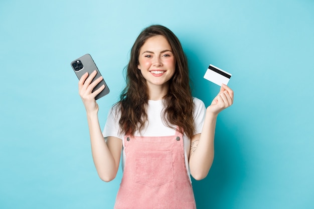 Smiling cute girl showing plastic credit card and smartphone, order something in store via mobile app, shop online, or pay for order, standing against blue background