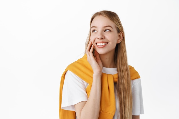 Smiling cute girl touching clean fresh skin cheek looking at upper left corner logo gazing at product banner with happy face standing over white background