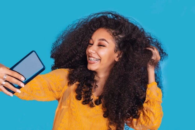 Smiling cute girl with curly hair takes selfie with a phone in her hands on a cyan background