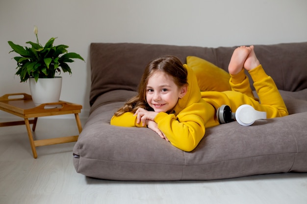 Smiling cute little girl in yellow clothes lying in bed with a headphones on listening to the music