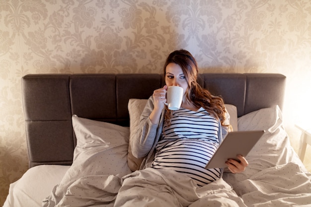 Улыбаясь милая беременная кавказская женщина с длинными каштановыми волосами, лежа в постели, пьет чай и читает о младенцах на планшете. Вечернее время.