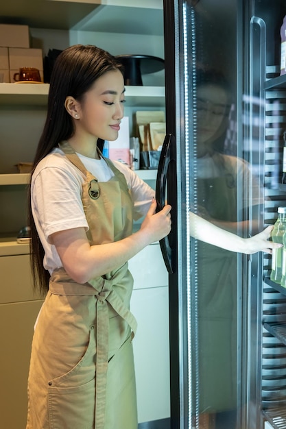 Photo smiling cute young longhaired asian woman reaching for a bottle in the refrigerated display cabinet