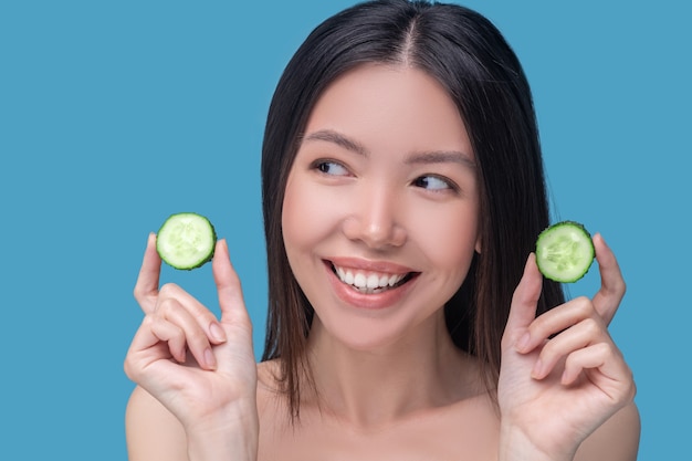 Smiling cute young woman holdingslices of cucumber